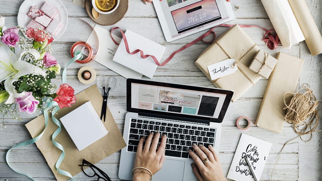 Prikaz drvenog stola iz ptičje perspektive na kojem su pokloni umotani u papir, ukrasne trake, buket cveća i na sredini su ruke koje kucaju na laptopu.