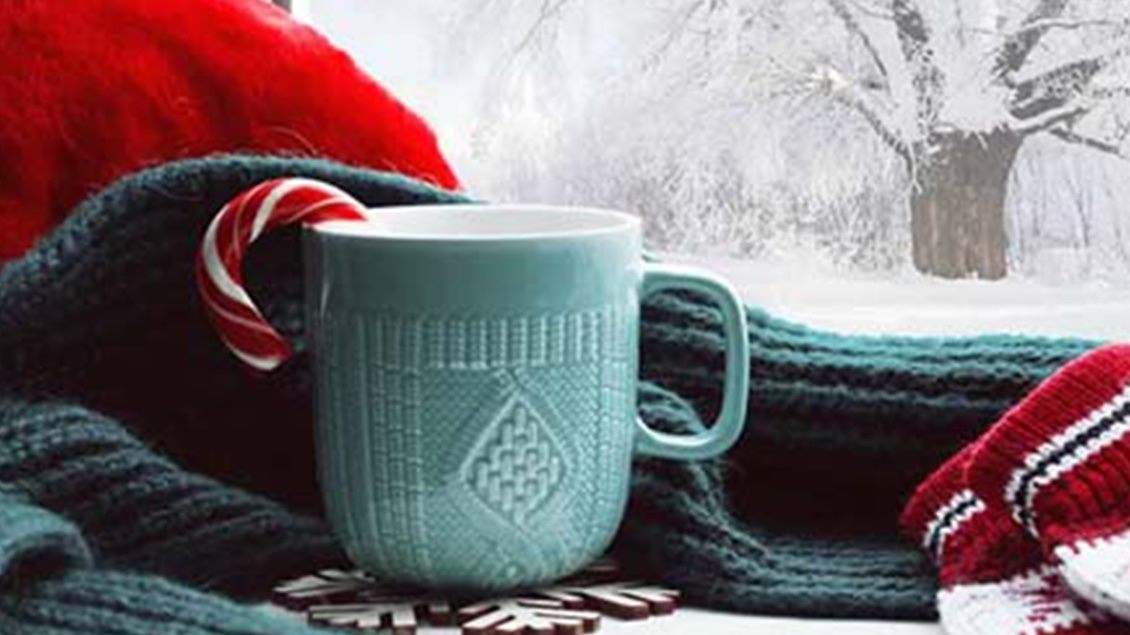 Prikaz zimske idile sa šoljom toplog napitka pored prozora sa pogledom na zavejani proplanak.