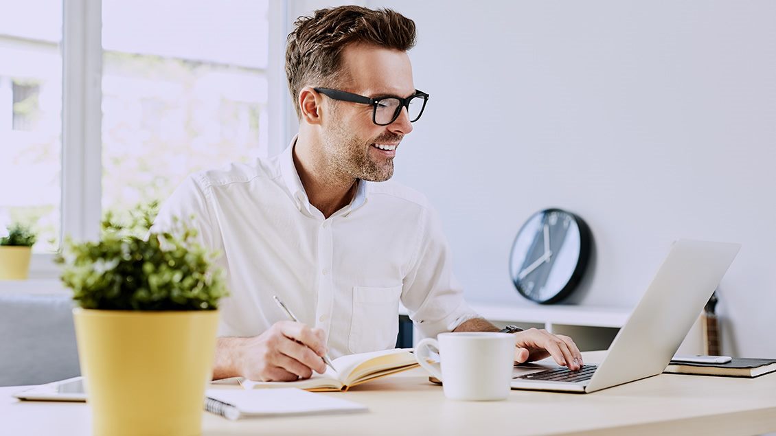 Nasmejan muškarac u beloj košulji čita mail-ove u kancelariji, sa sveskom i šoljom kafe na stolu.