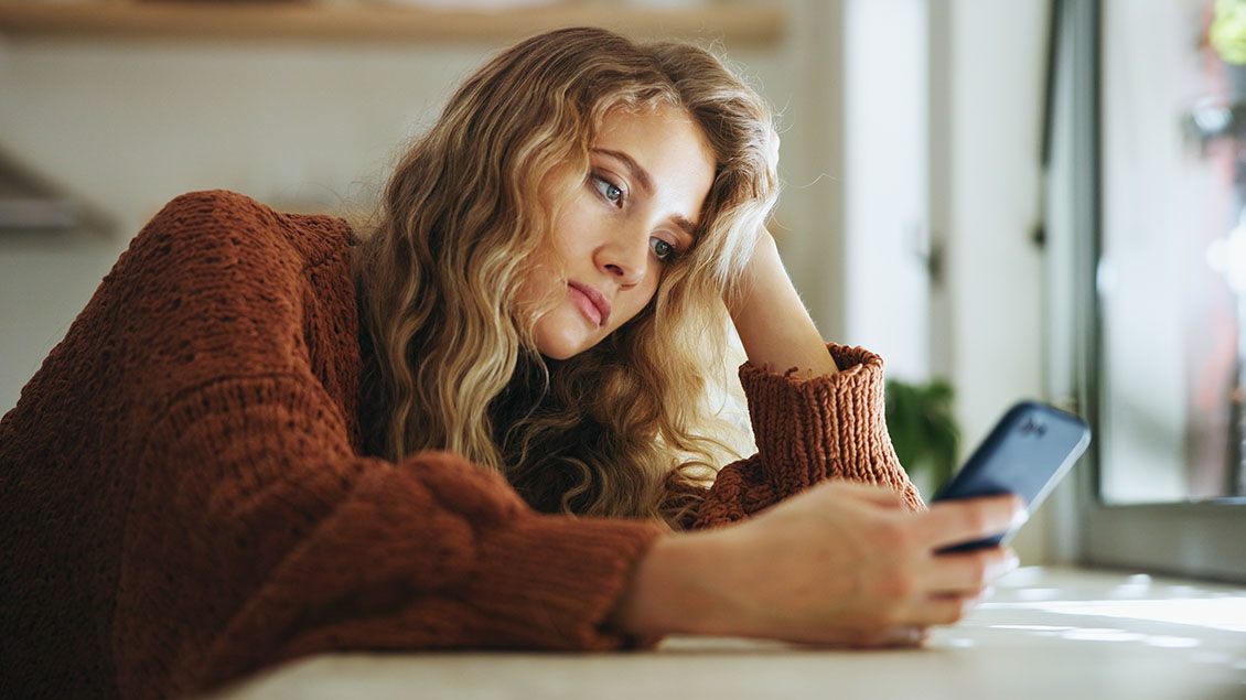 Žena zamišljeno gleda u telefon dok sedi za stolom