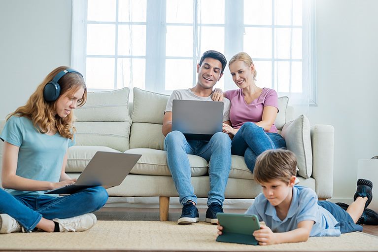 Porodica u dnevnoj sobi koristi Wi-Fi – roditelji na laptopu, tinejdžerka sa slušalicama i laptopom, dete sa tabletom.