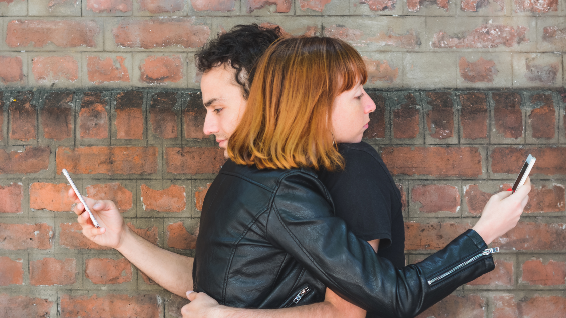 Dvoje mladih ljudi stoji zagrljeno, ali oboje gledaju u svoje mobilne telefone umesto jedno u drugo.